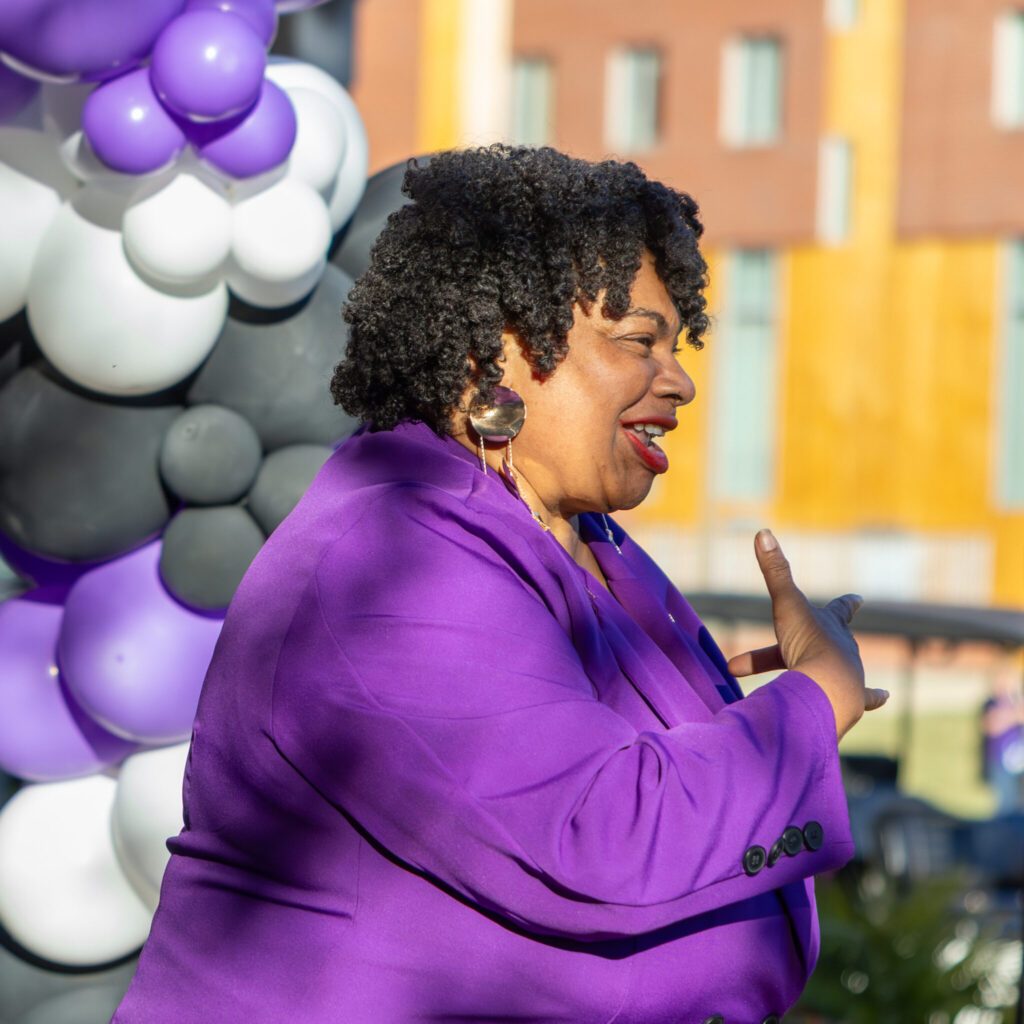A photo of a person in a bright purple pantsuit signing outdoors toward an unseen audience to the right.