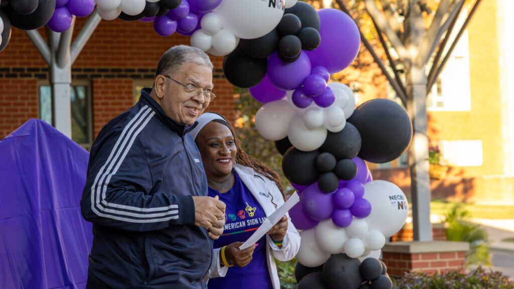 A photo of two people on a small outdoor stage addressing an unseen audience to the right. In the background is an arc of purple, black, and white balloons. The person on the right is holding a paper for the person on the left to follow.