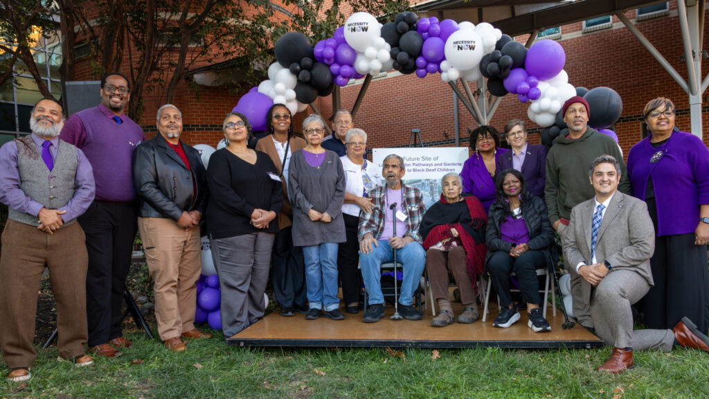 A photo of 16 people posing around a small outdoor stage, smiling for the camera. The stage has a decorative arc of purple, white, and blue balloons. Three people on the stage are seated. The rest stand around and on the stage, other than one kneeling individual.