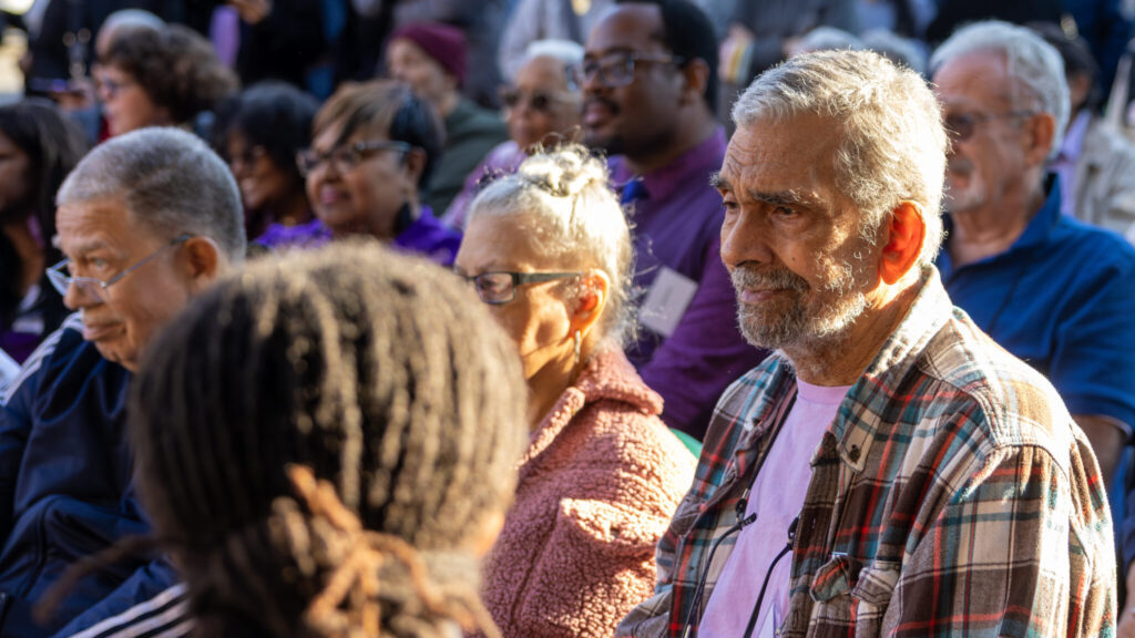 A photo of an older person framed centrally amongst seated people, outdoors in the shade on a sunny day. They are looking an unseen presentation to the left.