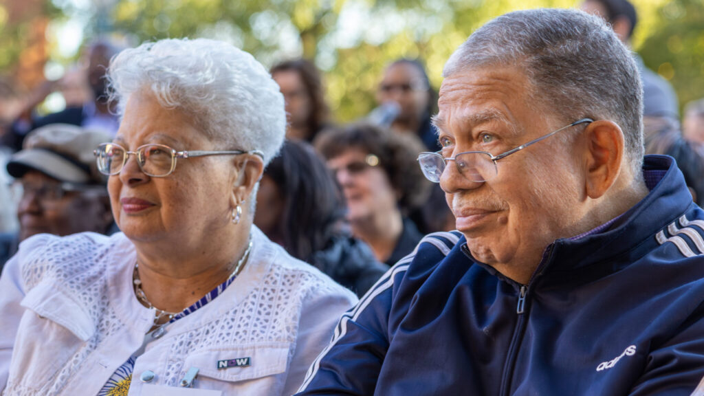 A closeup photo of two older people seated in an outdoor crowd. They are looking attentively toward an unseen presentation to the left. Both have glasses and graying hair.
