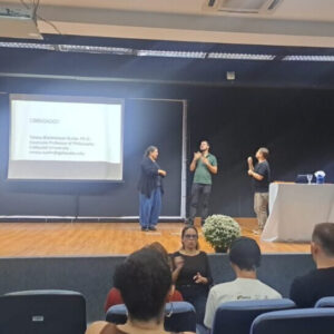 A photo of three people onstage giving a presentation. in sign language. They are facing each other and signing. A slideshow is projected on the left. There are various people in the audience, including a sign language interpreter.