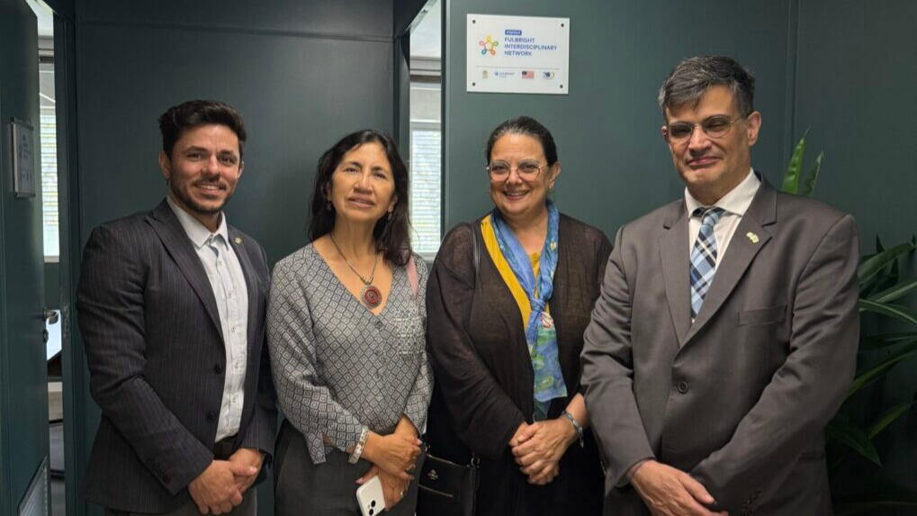 A photo of four people of varying identities in business attire, smiling for the camera. A small sign in the upper middle says "Fulbright Interdisciplinary Network".