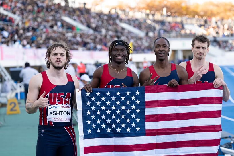 A photo of four track athletes posing for the camera, holding up a USA flag. They wear tight tank tops with red and dark blue that read "USA". Their free hands make various signs.