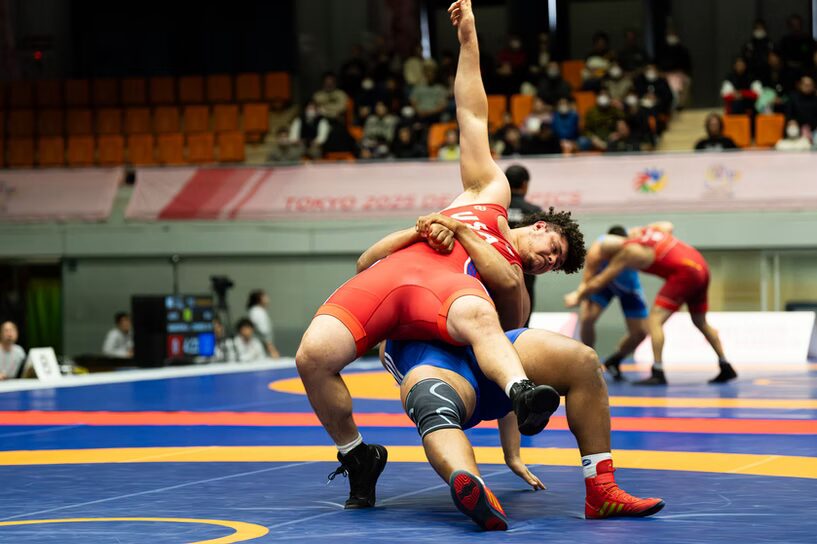 A photo of wrestlers in action. A wrestler in a red uniform reading "USA" is being pulled down from behind by a wrestler in a blue uniform. In the background are two other wrestlers, with bleacher seats in the distance.