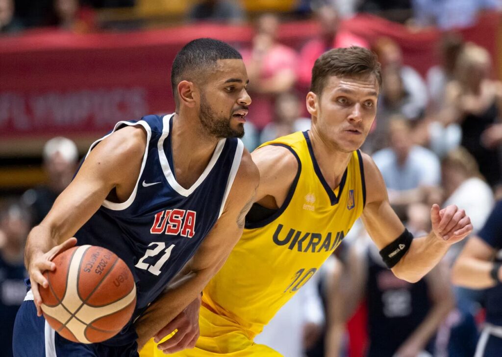 A photo of two basketball players running to the right. The player on the left is dribbling a ball. Their #21 dark blue jersey says "USA". The player on the right is defending and looking ahead. Their #10 yellow jersey says "Ukraine".