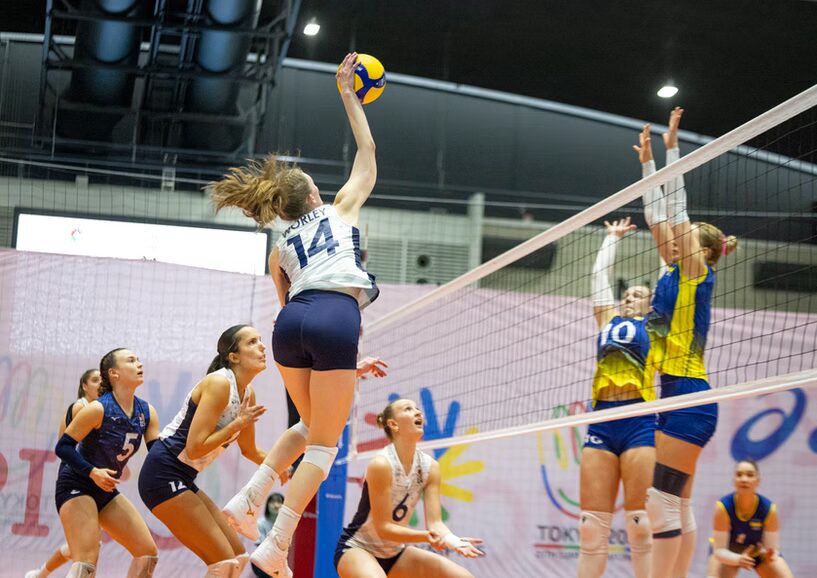 A photo of a volleyball spike in action. On the left, five players are visible with white and dark blue uniforms. In the foreground, #14 jumps and strikes a ball above the net. On the right, two players in blue and yellow uniforms jump by the net with arms raised to block. Another teammate is in the background watching.