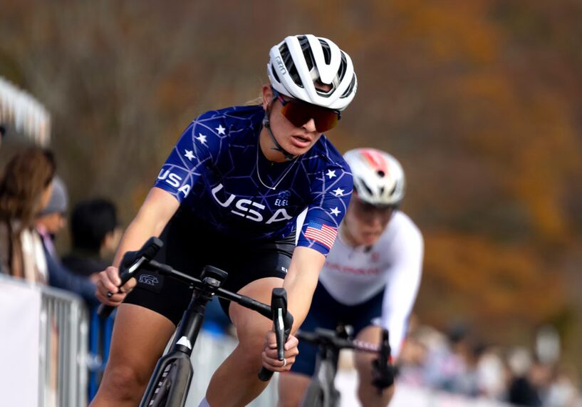 A photo of a cyclist in blue, black and white. Their jersey has "USA" on the chest and right sleeve, and a USA flag on the left sleeve. They are rounding a turn to their left.