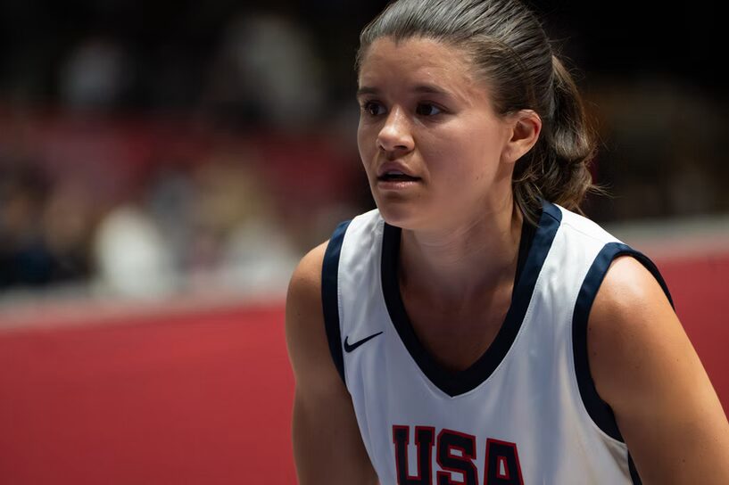 A photo of a basketball player looking attentively to the left. They wear a white uniform with "USA" in dark blue. Out-of-focus in the background are arena bleachers.