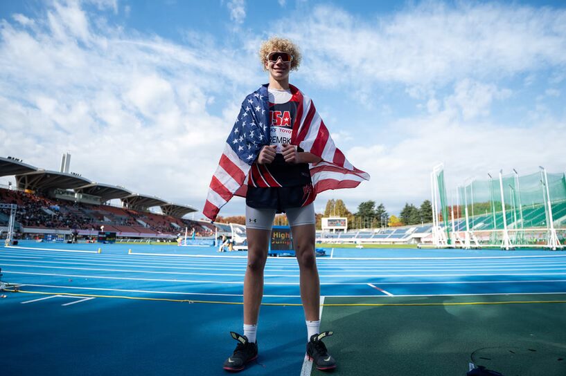 A photo looking up at a smiling track athlete with the USA flag wrapped around them. They stand on the sideline of a blue track in a stadium. They have curly blonde hair and dark glasses. They wear a dark blue and red uniform reading "USA".