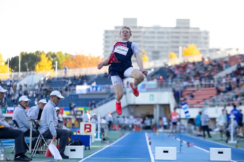 A photo of an athlete in mid-air during a long jump. They wear a dark blue and red uniform reading "USA". Their jump is on a track with judges watching. A crowd is out of focus in the background.