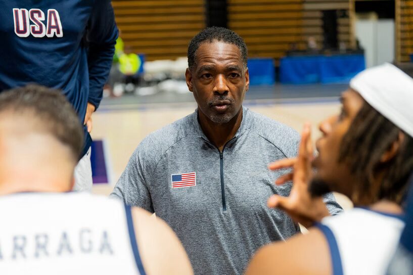 A photo of a coach addressing a basketball team huddle. They are wearing a gray pullover with a USA flag on the right chest. Out of focus in the foreground are two players in white. In the background, top left of the image, is a person in a dark blue shirt that says "USA".