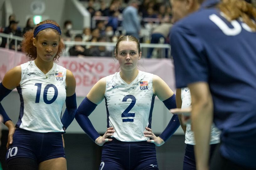 A photo of two volleyball players attentively watching an out-of-focus coach in the foreground on the right. The players wear white jerseys with a USA flag, dark blue shorts, and dark blue arm sleeves. Out of focus in the background are bleachers, with a banner hanging over the front that says "Tokyo".