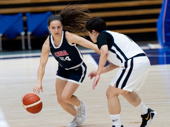 A photo of two basketball players on a court. The player on the left, wearing a dark blue uniform with "USA", is dribbling forward while the player on the right in white defends.
