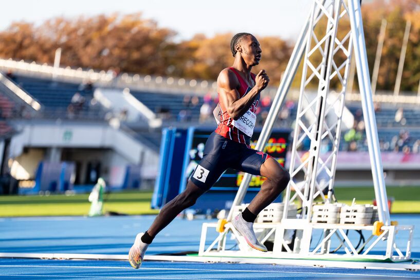 A photo from the side of a runner mid-stride on a blue track going to the right. They wear a dark blue and red uniform. In the background are track decorations: a metal tower, scoreboard, and bleacher seats.