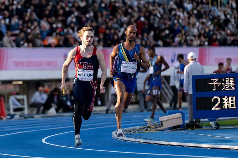 A photo of two runners in mid-stride rounding the turn of a blue track. The runner in front is wearing dark blue and red reading "USA", and holding a baton in their right hand. The runner in back has a colorful uniform and baton in their right hand. In the far background is a crowd in bleachers.