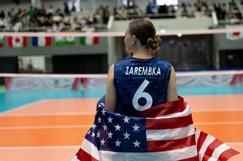 A photo of a volleyball player looking away from the camera with an out-of-focus volleyball court in the background. They wear a dark blue jersey with a white name and number: "Zarembka #6". A USA flag is wrapped around their midsection. Past the volleyball court bleachers with a crowd are visible.