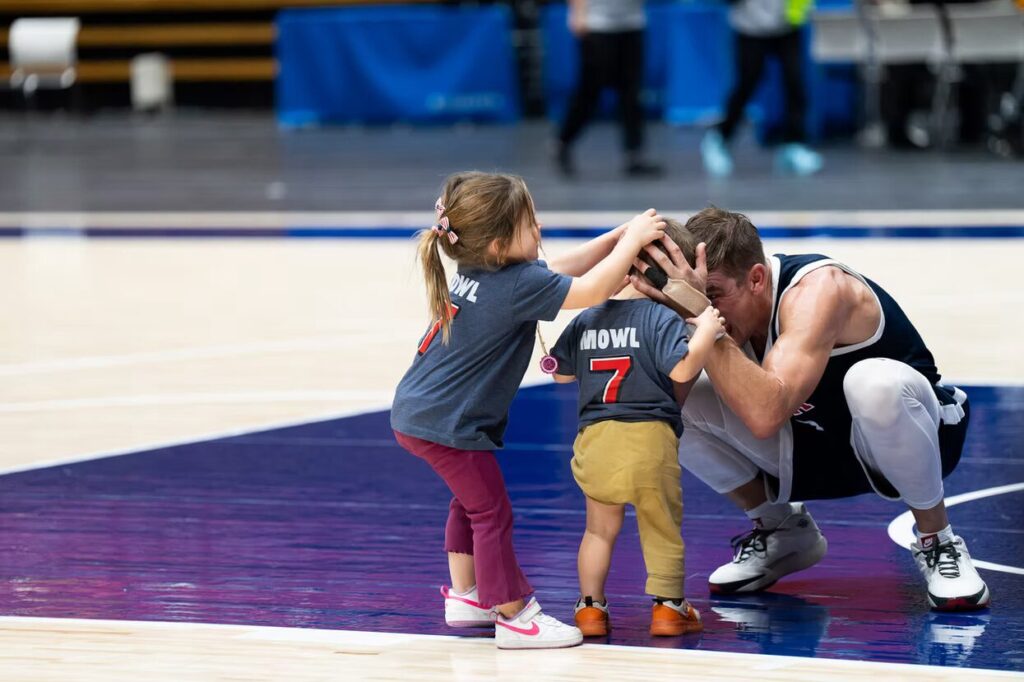 A photo of a basketball player squatting to engage with two small children who have t-shirts that resemble jerseys with "Mowl" and the number 7. The player is nuzzling the closest child playfully, while the other child paws at the nuzzle.