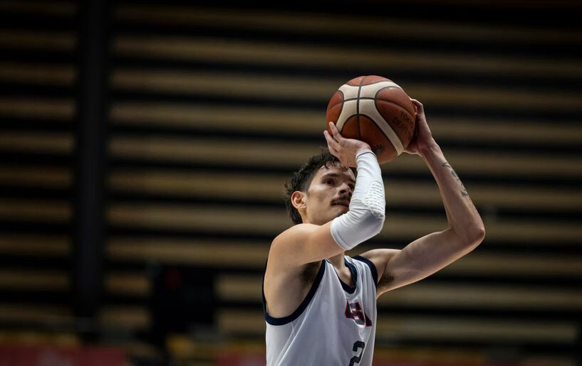 A photo looking up at a basketball player shooting a free throw. They wear a white jersey that can be assuming to read "USA" and a white elbow sleeve.