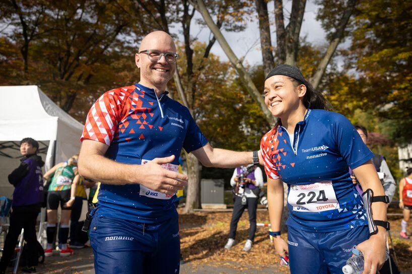 A photo of two orienteers smiling in a park. They are wearing uniforms of dark blue with a red and white splash over the right shoulder. They have numbers pinned over their stomaches. In the background, various people are milling.