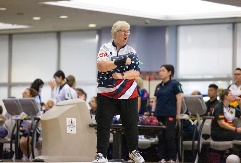 A photo of a bowler celebrating with a yell and flex in a busy bowling alley. They are wearing a gaudy bowling shirt with USA colors and styling. They have white hair and glasses.