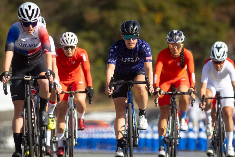 A photo of six cyclists biking toward the camera. Prominent in the middle is a cyclist with a jersey that says USA on the chest. They have a black helmet and dark googles.