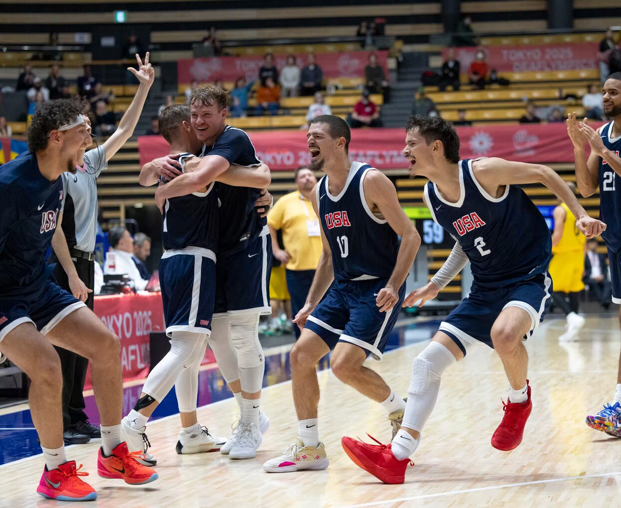 A photo of six basketball players running toward each other and celebrating on a court. They wear dark blue jerseys that say "USA". In the foreground, two players in the center right of the image are mid-landing and cheering towards their bench. In the center left, two other players are smiling and hugging. On the outsides of the image, two other players join the celebration.