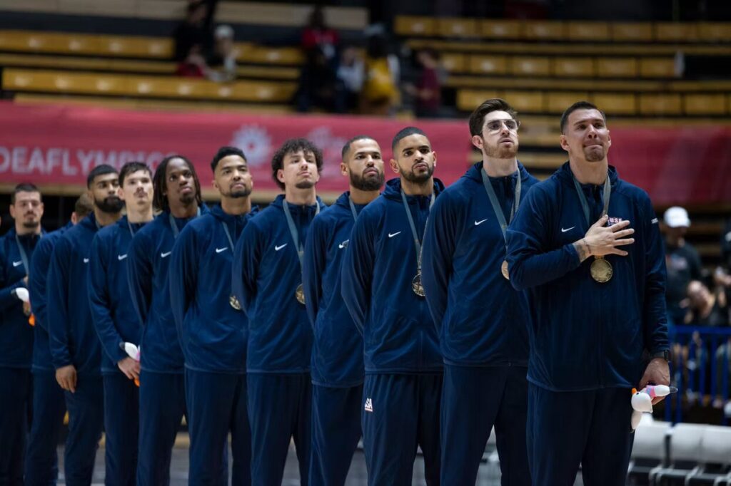 A photo of eleven athletes in dark blue jumpsuits lined up with gold medals around their neck. They are looking at a flag offscreen to the upper right in an arena.