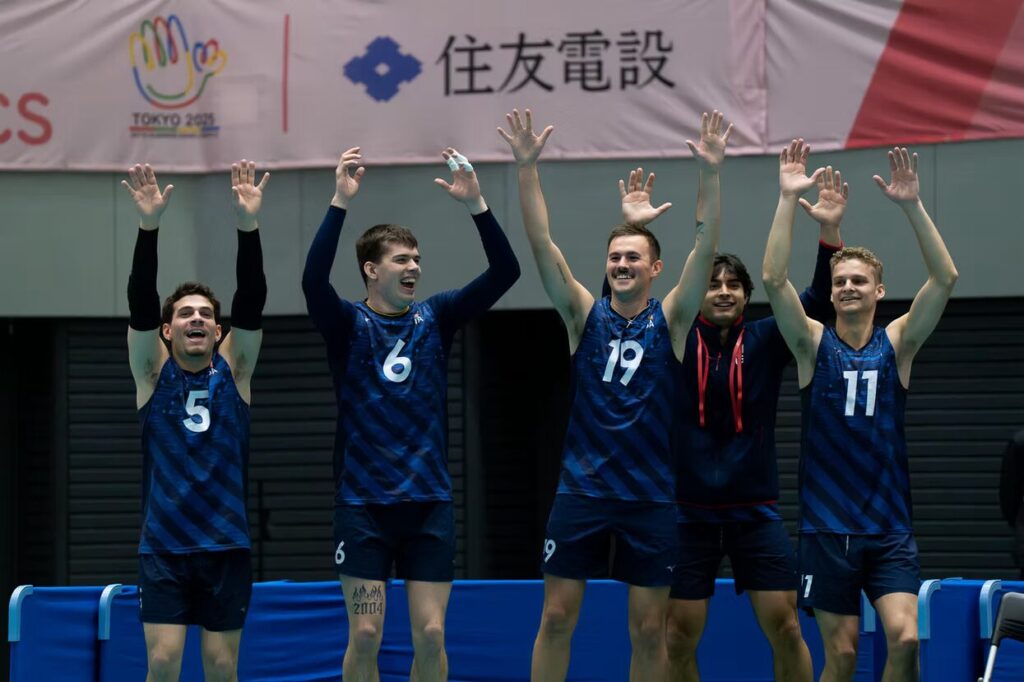 A photo of five volleyball players smiling and raising their arms up on the sideline. They are wearing dark blue and black striped jerseys. Four numbers are visible: 5, 6, 19, and 11. In the upper background is a banner with "Tokyo" and some Japanese kanji.