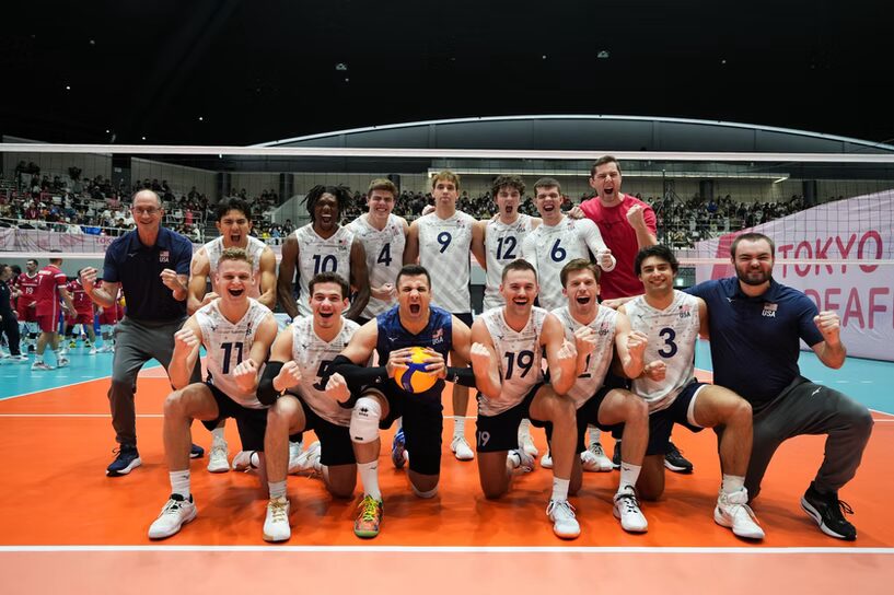 A photo of 12 volleyball players and three coaches posing happily for the camera on a volleyball court in front of the net. 11 players are wearing white and gray striped jerseys with dark blue numbers. The other player has a dark blue jersey. In the right background a wall with "Tokyo Deaf" is visible.