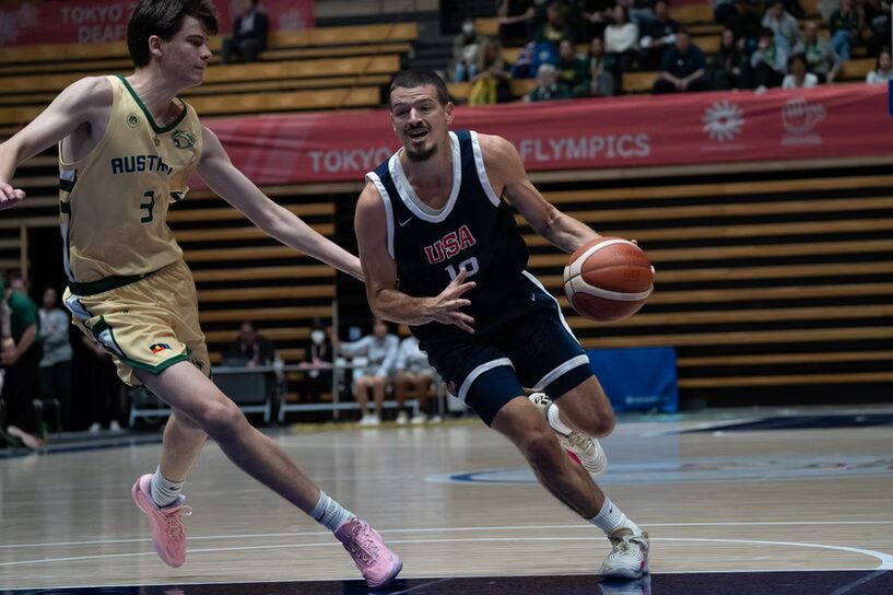 A photo of two basketball players. In the center, a player with a dark blue jersey reading "USA" drives intensely with the basketball. On the left, a defender with a gold jersey reading "Austria" tries to keep up.
