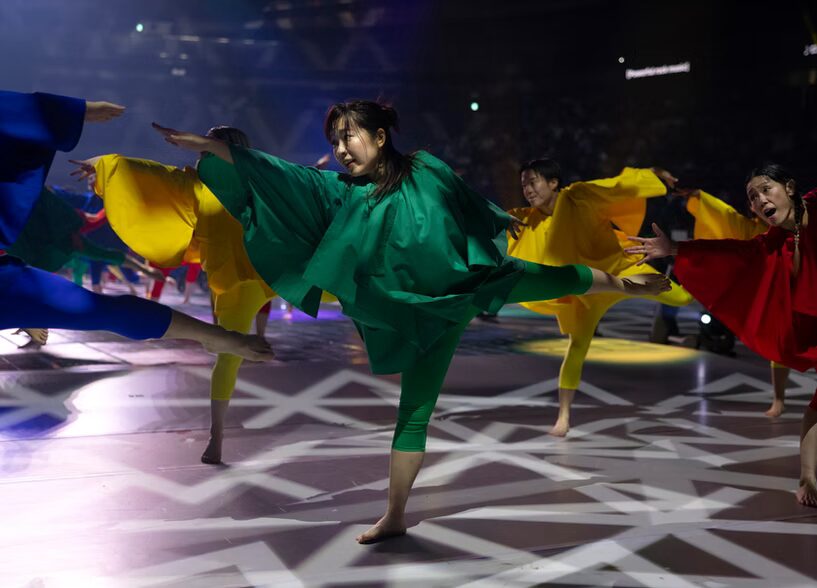 A photo of five dancers in bright, solid-color outfits on the floor of a dark arena. They are posing into a T shape, with their right arm and left leg straight off the ground. In the center, the most prominent dancer wears green. Behind, two wear yellow. Blue and red are also visible. Many more dancers are obscured in the background.