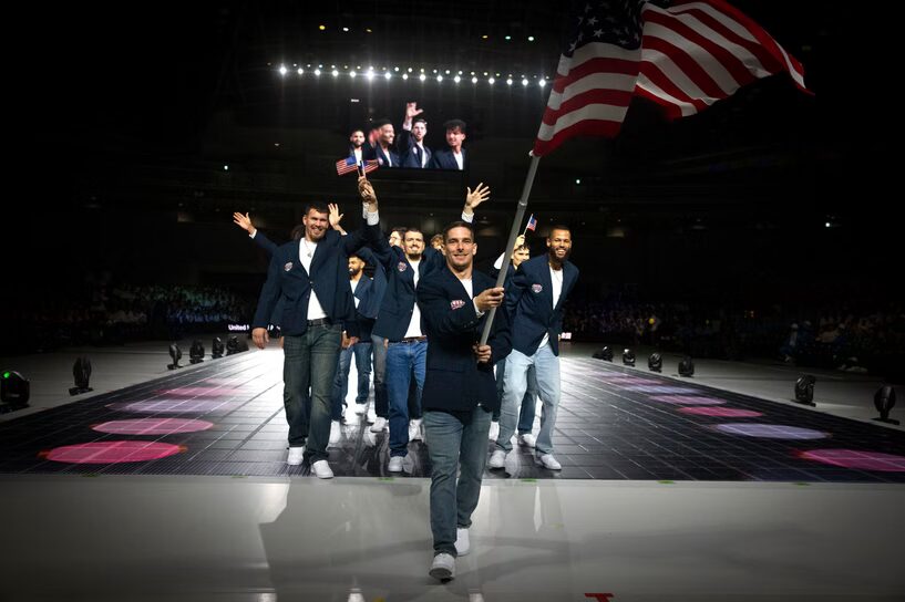 A photo of a team of USA athletes walking toward the camera, on the floor of a dark arena. It is clearly the front of a line. They are wearing dark blue blazers and white shirts. One person in front is holding and waving a large USA flag. In the upper background is a large screen showing a video camera's view.