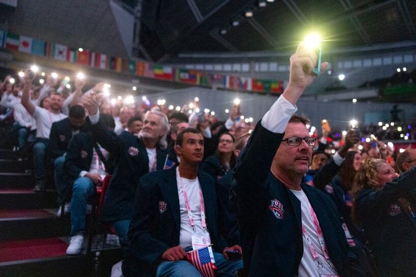 A photo looking up at seats in an arena with many people holding up their smartphones with flashlights on. The people in the photo are wearing dark blue blazers with white shirts. They appear to be athletes for Team USA.