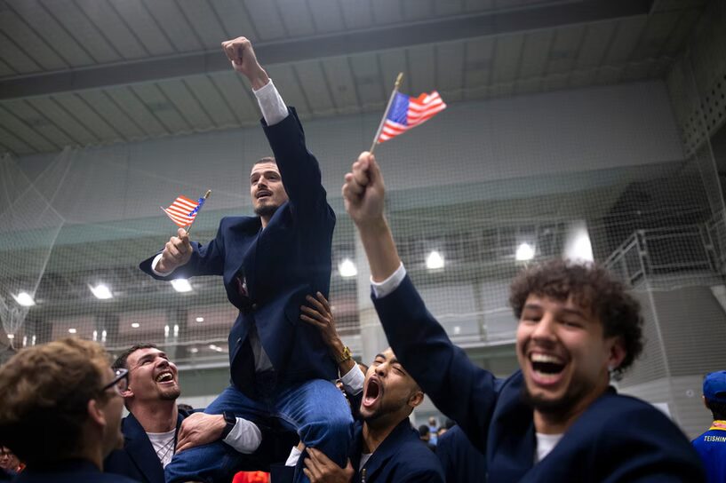 A photo of a person in a dark blue blazer and white shirt being held on the shoulders of two similarly-dressed teammates. They are grasping a small USA flag and raising their left arm. In the foreground, another teammate is cheering the camera with a flag.