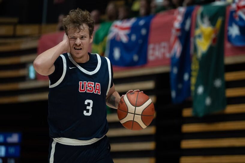 A photo of a basketball player dribbling with their left hand. They wear a #3 dark blue jersey reading "USA" over a black sleeveless shirt. Their right hand is over their right ear and their right eye is winking, either in pain or to signal a play to their teammates.