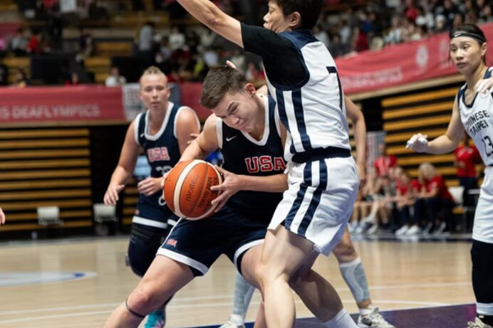 A photo of four basketball players on a court in an arena. Prominent in the foreground, two players jostle under the hoop. The player on the left is wearing a dark blue uniform with "USA". They have the ball and push their left shoulder into the player on the right, who has their hands up. They wear a white uniform.