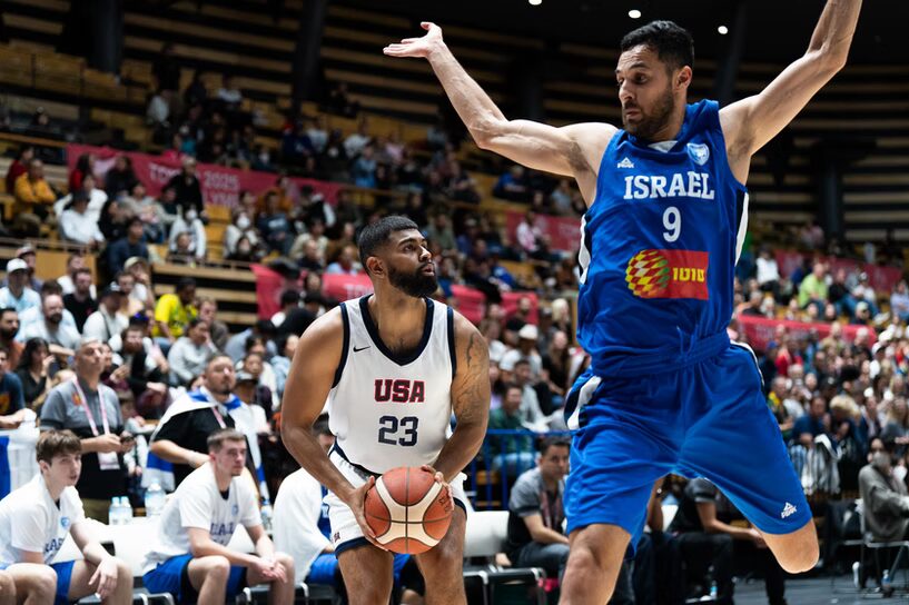 A photo of two basketball players with a crowd in the background. The player on the left is crouching to begin a jump shot with the ball. They wear a #23 white jersey reading "USA". On the right, their defender jumps in front with flailing arms. They wear a #9 blue jersey reading "Israel".
