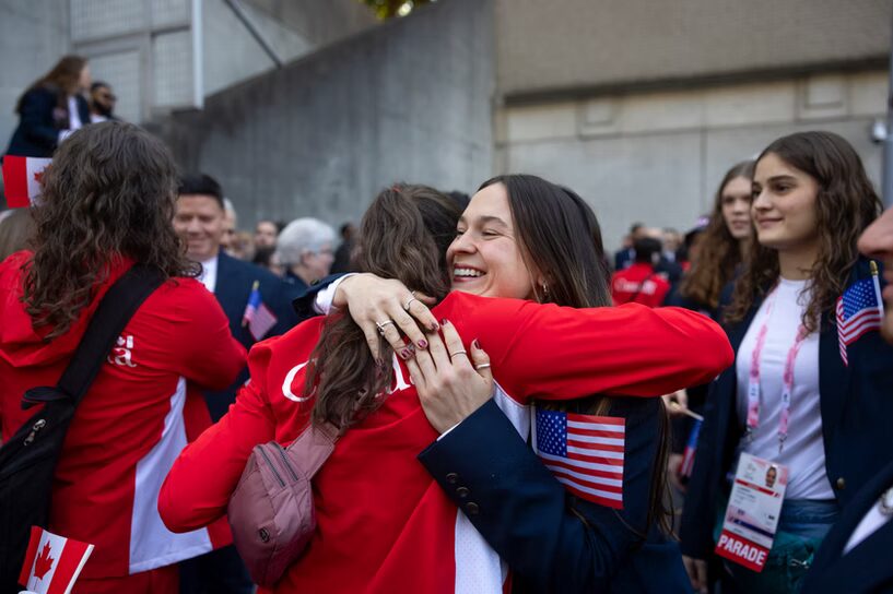 A photo of two people hugging. There are similarly-dressed people behind each person. To the right are dark blue blazers. To the left are bright red and white sports jackets. It can be assumed the huggers are athletes on Team Canada (left) and Team USA (right). The USA hugger is smiling brightly.