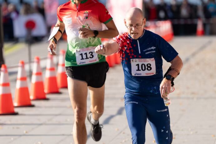 A photo of two orienteers crossing a finish line. Prominent in front is an orienteer with a dark blue uniform and the number 108 pinned over their stomach. They are visibly tired. In the background is a line of orange cones.