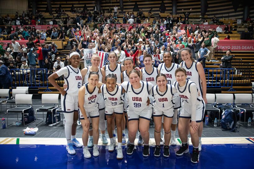 A photo of twelve basketball players smiling and posing in front of arena bleachers full of supporters. The players wear white jerseys reading "USA". Two USA flags are visible in the crowd.