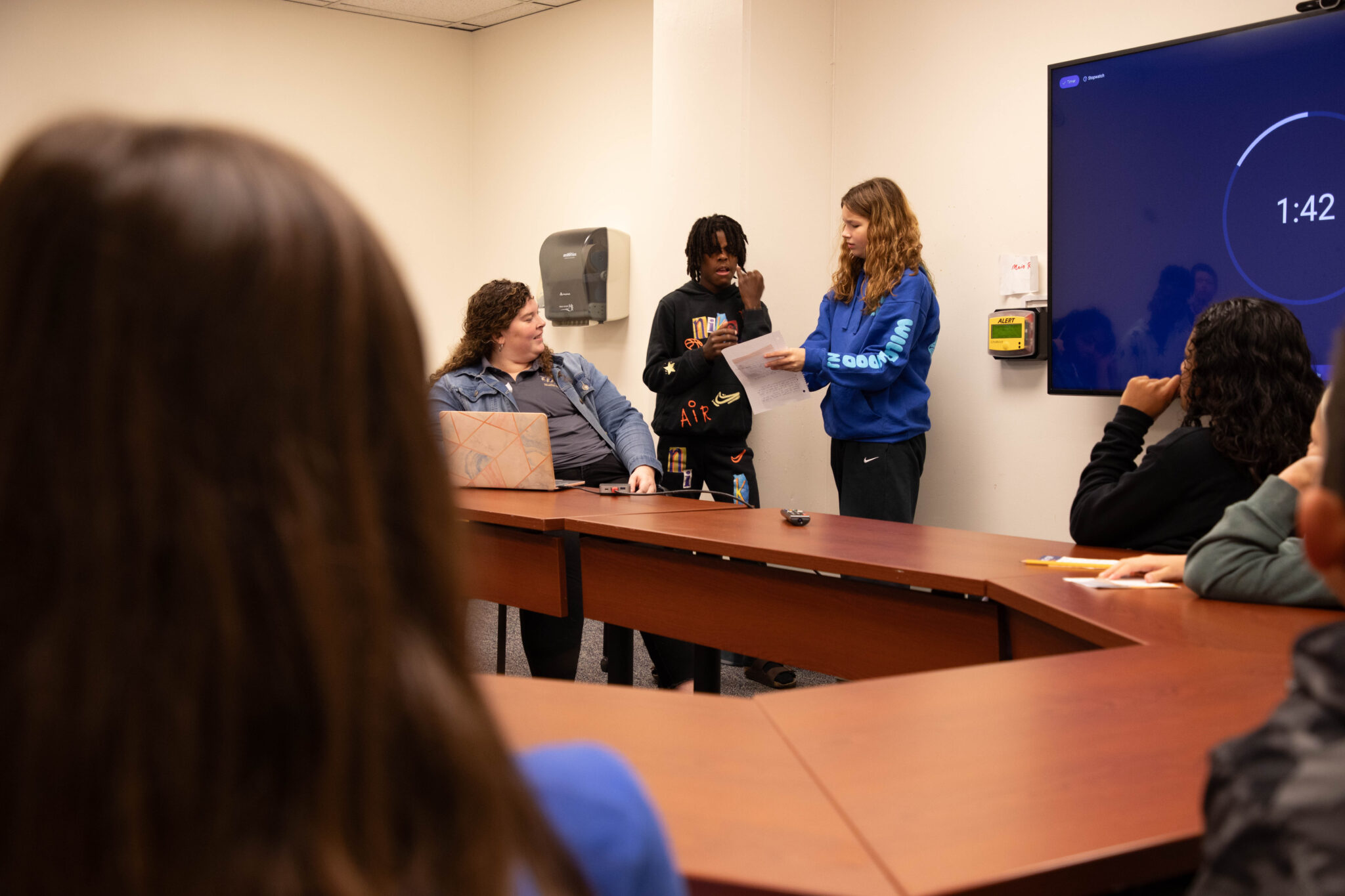 People are sitting around a conference style table. We see the back of one person's head and then across to a woman seated with a computer in front of her. Next to her are a boy and girl who are standing and discussing something.