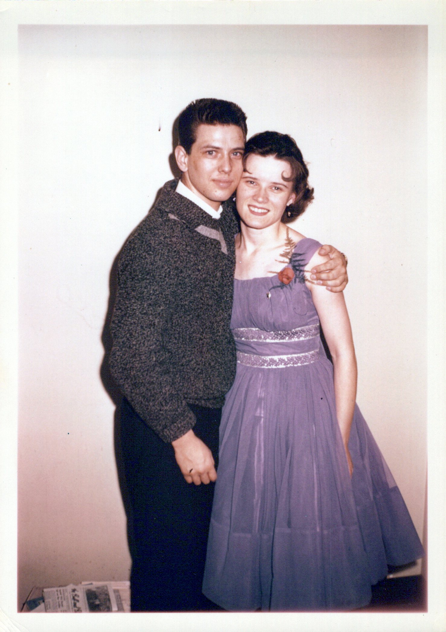 A young man and woman stand next to each other with the man's arm around the woman's shoulder. He wears a sweater and slacks and she is in a purple dress.