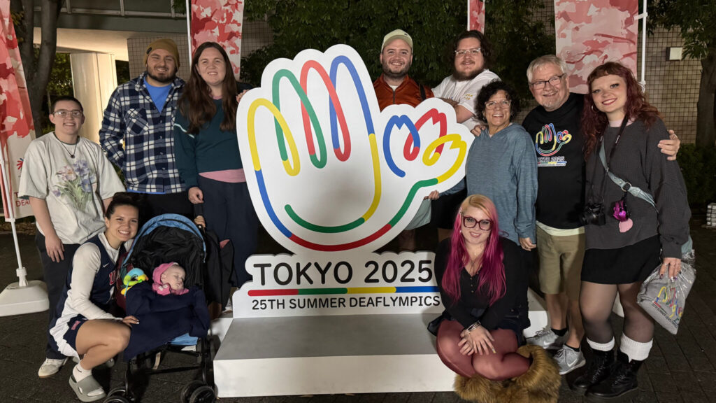 A photo of 11 family members smiling for the camera. They are on either side of a large sign with a colorful logo and text: "Tokyo 2025 / 25th Summer Deaflympics".