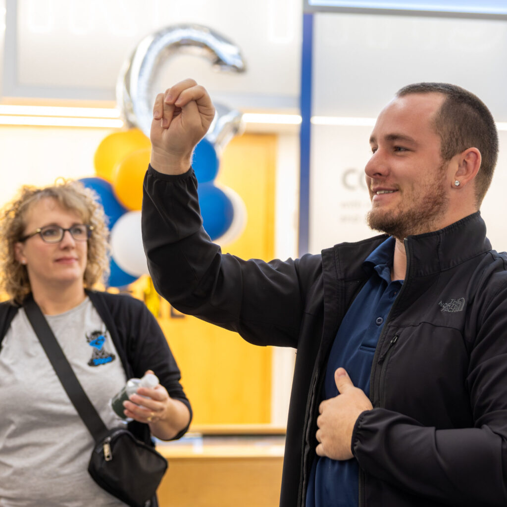 A photo of a tour leader signing to an unseen group to the left. They are wearing a black jacket over a blue shirt. On the left of the image, in the background, one tour member is visible.