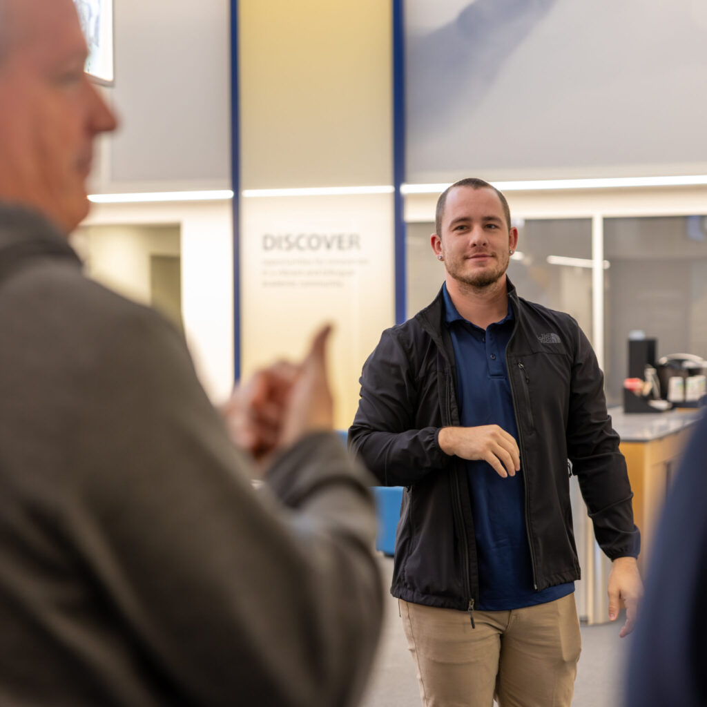 A photo of a person smiling and watching a signed conversation, which is out of focus in the foreground. The person is wearing a black jacket, blue shirt, and khaki pants.