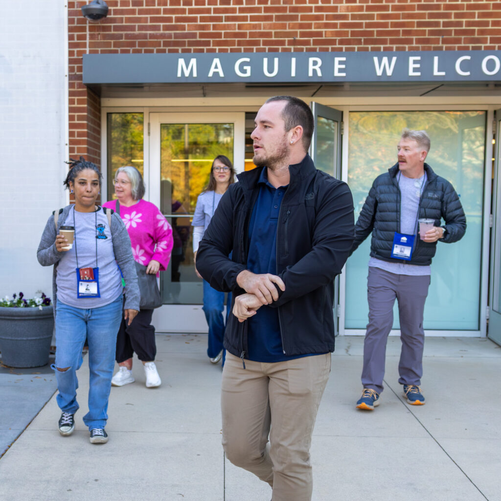 A photo of a tour group exiting a building. Prominent in the center foreground is the tour leader, looking to the left. They wear a black jacket, blue shirt, and khaki pants. Behind them are four group members looking around. Above the door they're leaving is obscured text: "Maguire Welcome—".