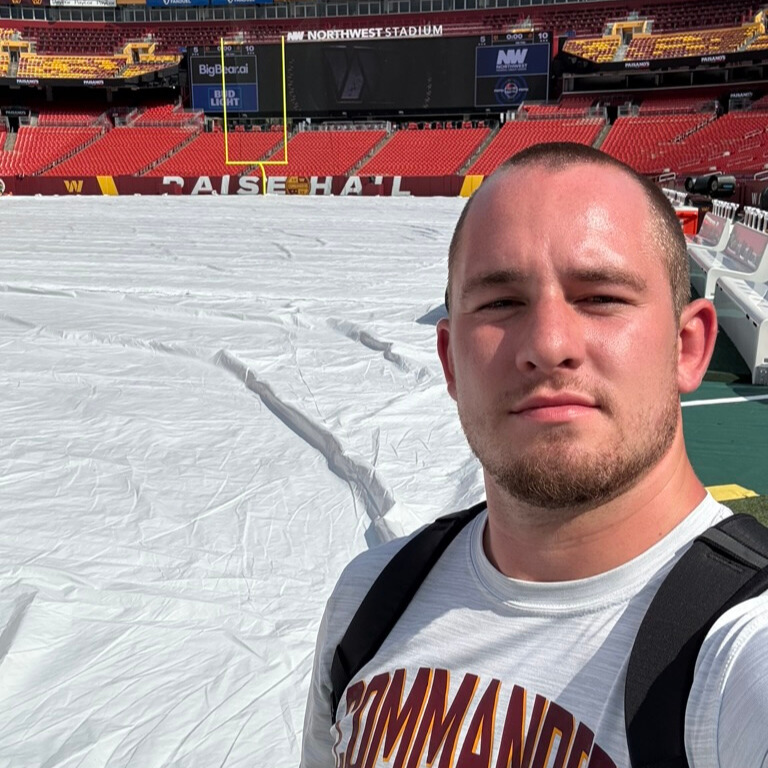 A photo of a young adult taking a selfie on the sideline of a football field. They are wearing a white Washington Commanders shirt. The field is covered with a tarp.