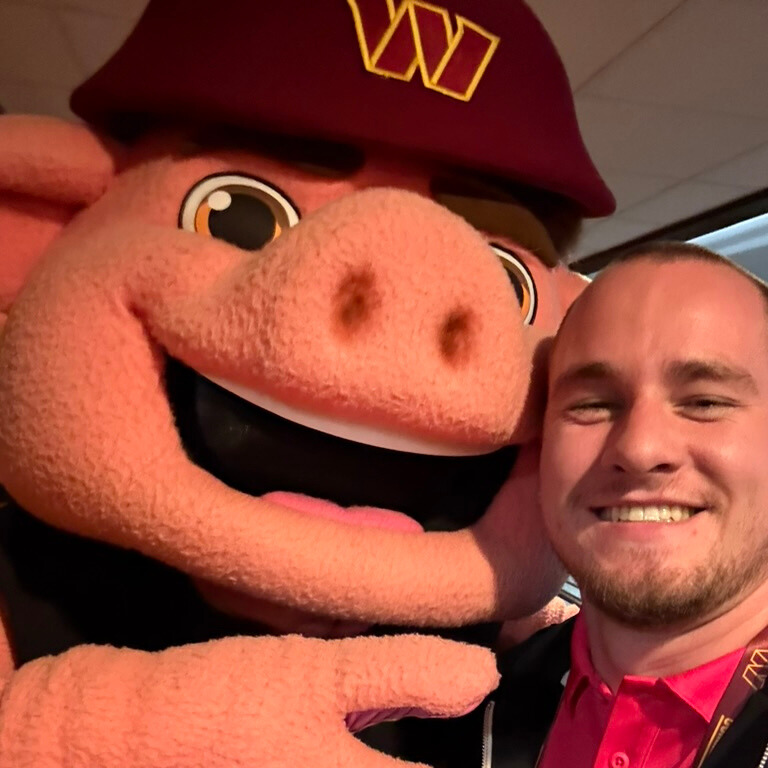 A photo of a person, right, smiling for a selfie with the Washington Commanders mascot, left. The mascot is an anthropomorphized pig with a Washington Commanders hat.