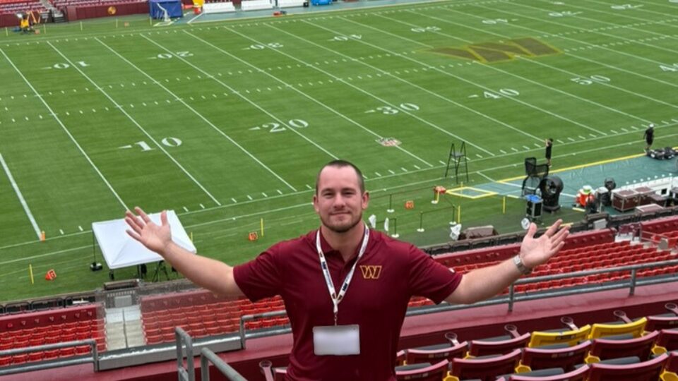 A photo of a young adult standing in the seats of an empty stadium, with a football field in the distance. They are holding their arms out to their sides. They wear a red polo with the Washington Commanders logo and a lanyard.